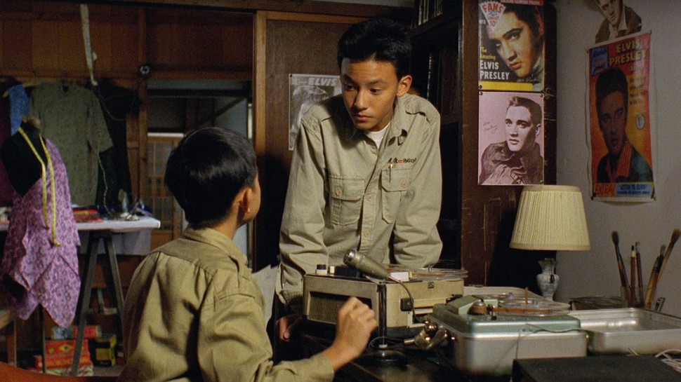 two young Taiwanese boys in military uniforms talk to each other over a tape recorder with Elvis posters in the backgroundalr