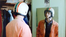 a young white man wearing a striped motorcycle helmet looks in the mirror